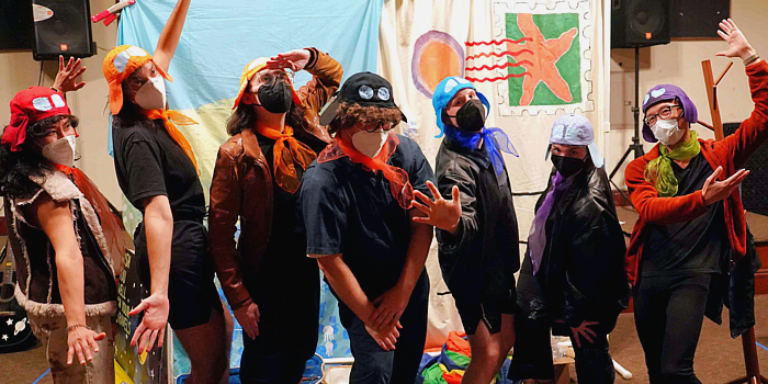 a photo of the cast of Greetings From in costume. Seven performers are standing in a row, posing dramatically. They all have KN-95 face masks. They each have a different colored fake pilot helmet and matching ascot. Several have leather jackets. Behind them is the backdrop which resembles a giant postcard.