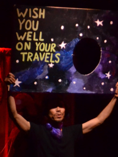 a close up of an Amelia whose face is in shadow, holding up a giant cardboard postcard with an outer space background which reads 'Wish You Well On Your Travels'