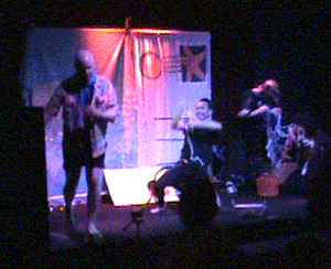 a moving clip from the play. In center stage, a performer playing a spider frantically pulls seemingly endless rope out of himself and throws it wildly into the air. To his right, a man in a Hawaiin shirt seems to be crying, to his left, two characters are playing with a bundle of rope woven into a net shape.