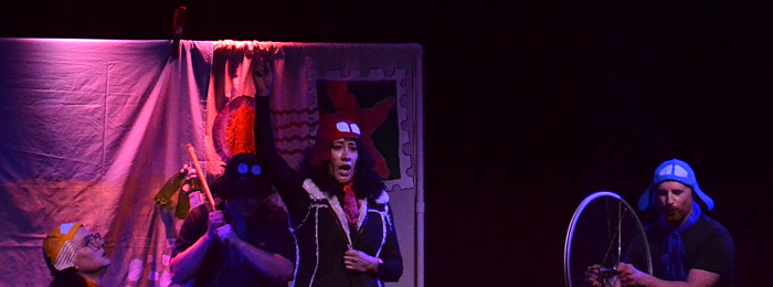an image captured from the play Greetings From. Four Amelias are seen, each one has a different colored hat resembling a pilot's helmet. One Amelia in a red hat and vest stands in front holding her arm up triumphantly. Two other Amelias crouched over are holding strange props - a stick with bottles attached, and a bicycle wheel. In the background is a large backdrop painted to look like a giant postcard.