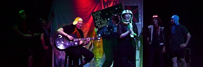 Six Amelias are on stage hanging out casually, one is holding up a giant postcard. A seventh Amelia is playing guitar. In center stage, a performer dressed in a space helmet is singing.